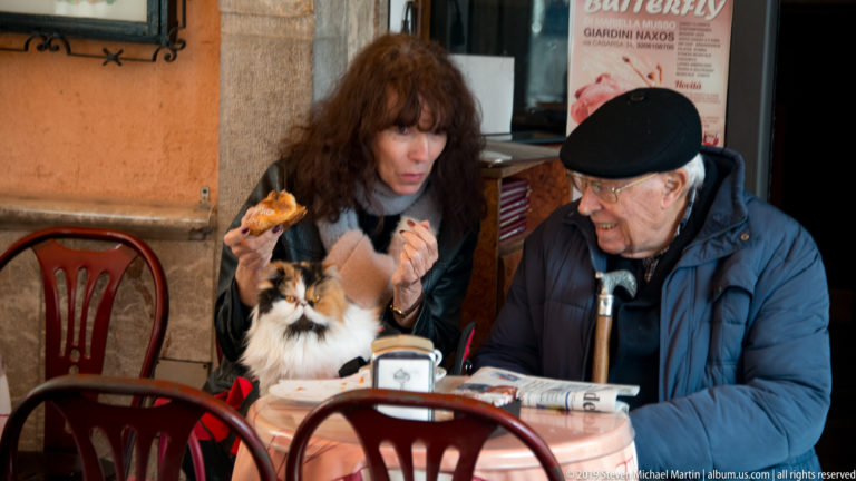 Two seated café patrons with a small long-haired dog looking up at food held over the table.