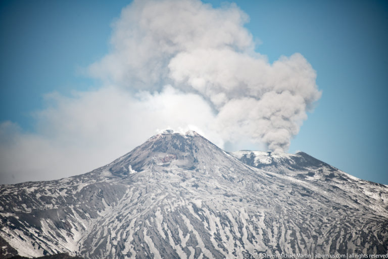 Snow-covered summit ridges of Mount Etna beneath a broad gray-white plume emerging from the crater area.