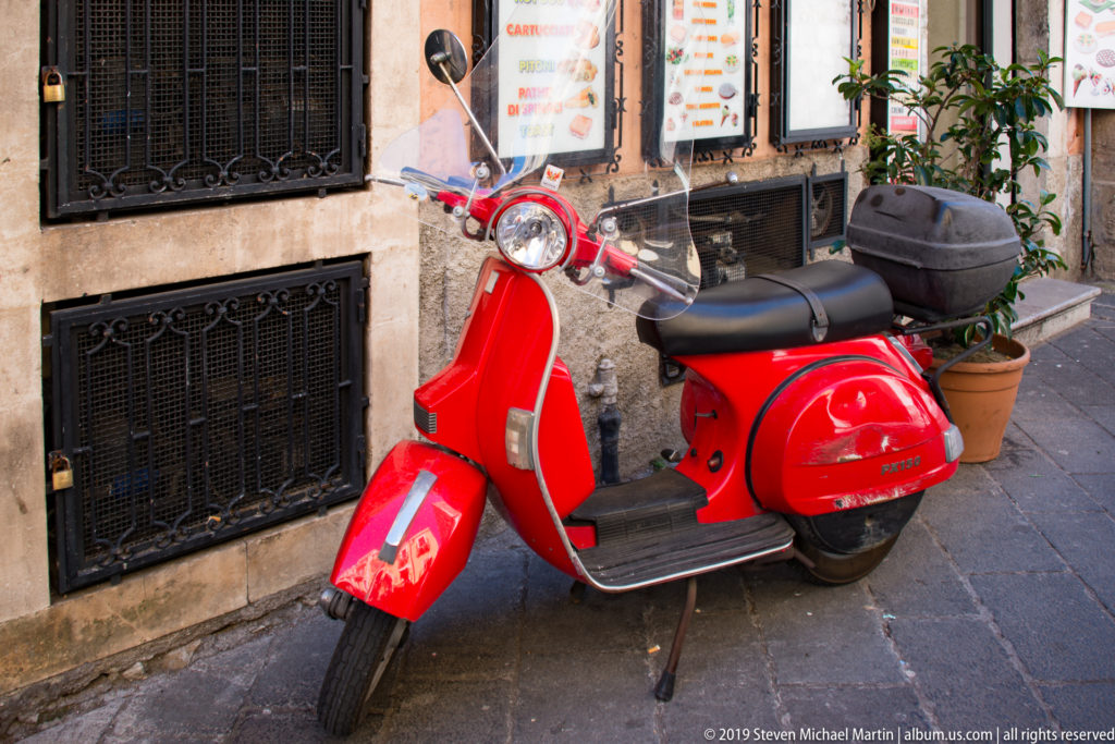 Bright red motor scooter with windshield and rear case parked beside restaurant menus on a narrow stone-paved street.