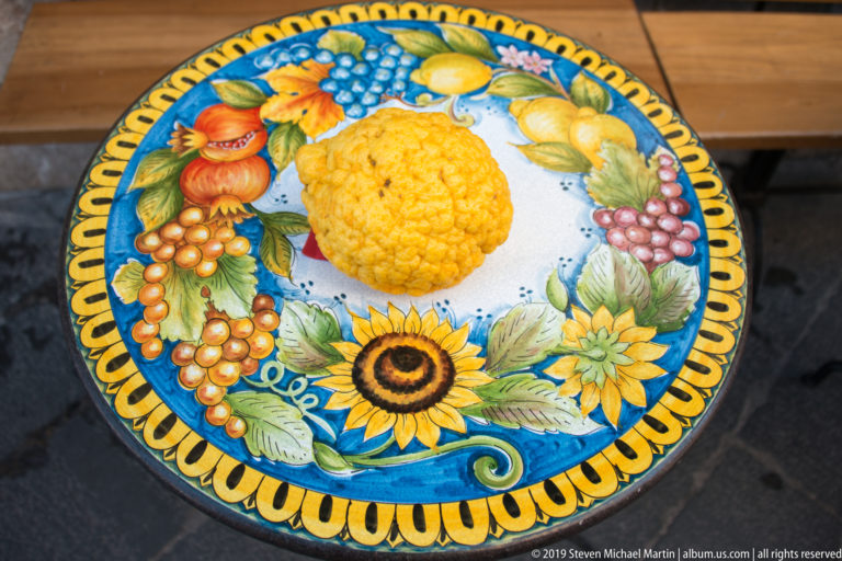 Round glazed ceramic table decorated with raised lemons, grapes, pomegranate, sunflower, leaves, and colorful fruit motifs.
