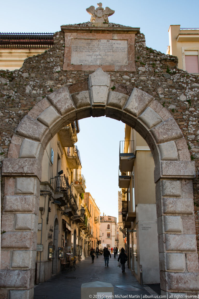 Large rusticated stone arch in an old wall opening onto a pedestrian street lined with balconies and shops.