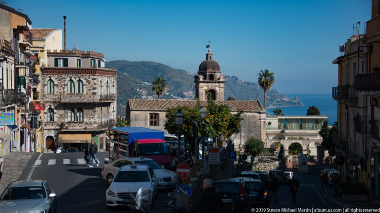 Busy descending street with cars and pedestrians, ornate buildings, a church bell tower, mountains, and blue sea beyond.