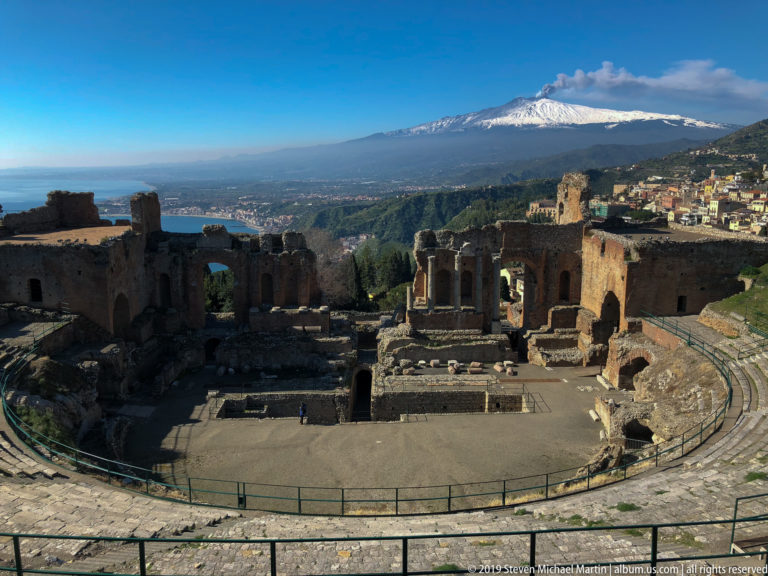 Ancient theatre stage ruins below a broad coastal landscape, with snow-covered Mount Etna dominating the horizon.