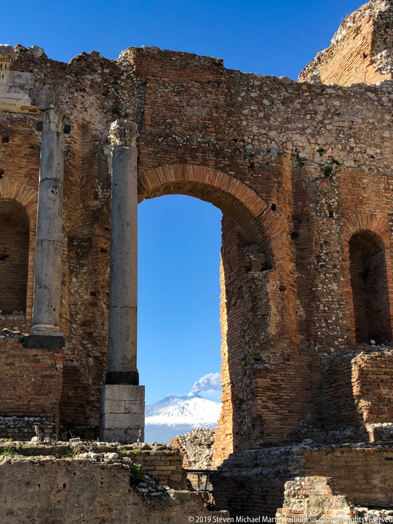Large brick arch and two pale columns in the ancient theatre framing snow-covered Mount Etna through the opening.