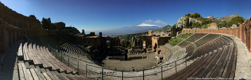 Ancient theatre seating divided between deep shade and sunlight, with stage ruins and Mount Etna in the distance.