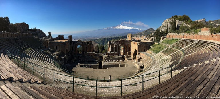 Semicircular ancient theatre seating around stage ruins, with snow-covered Mount Etna beyond under a clear blue sky.