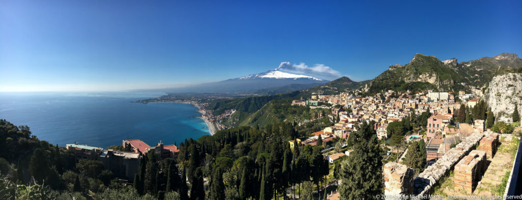 Taormina hillside and limestone cliffs above the blue coast, with snow-covered Mount Etna in the distance.