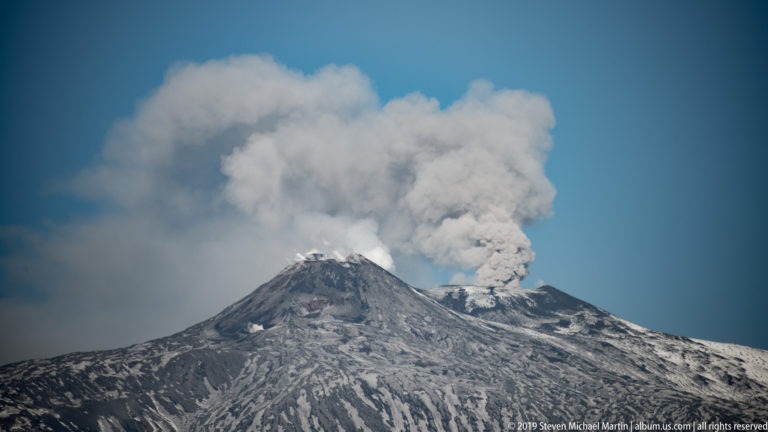 Close view of Mount Etna’s snow-streaked summit with a dense gray-white volcanic plume rising into blue sky.