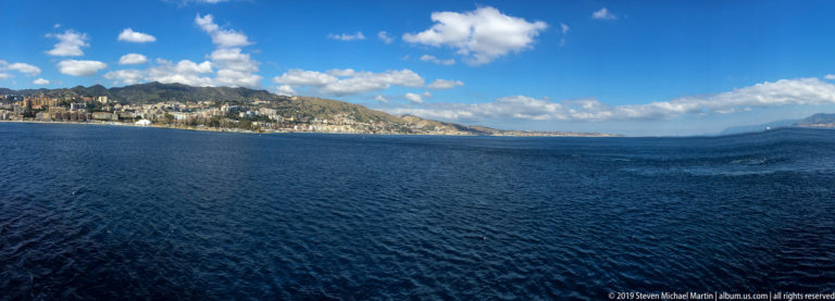 Wide blue strait with a hilly developed shoreline on the left and distant land beneath scattered clouds.
