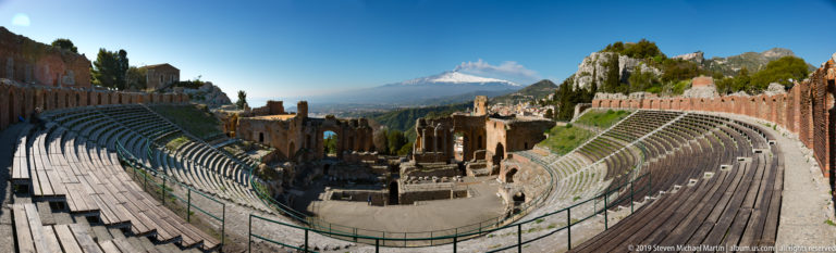 Curved seating tiers and stage ruins of the Ancient Theatre of Taormina with Mount Etna centered in the distance.