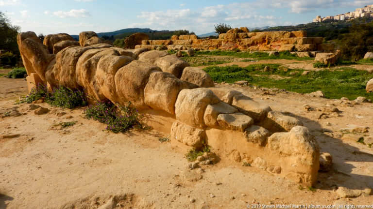 Side view of a colossal recumbent stone male figure lying among temple blocks with small flowers at its base.