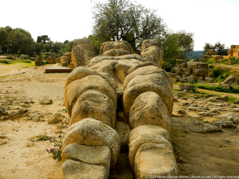 Foreshortened view from the feet of a colossal recumbent stone male figure lying among ruins and olive trees.