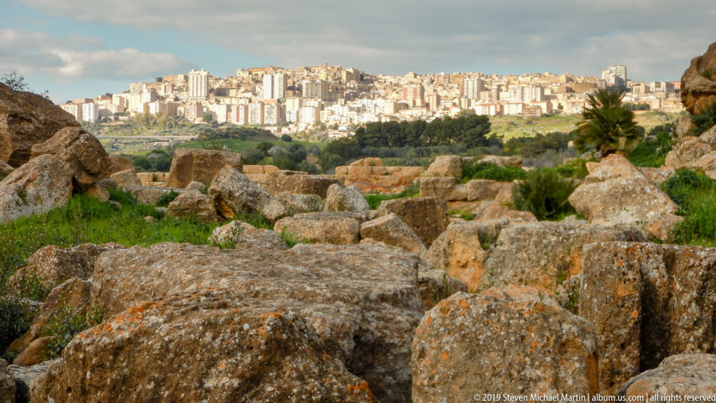 Massive weathered stone blocks in grass with modern Agrigento densely covering the distant ridge.