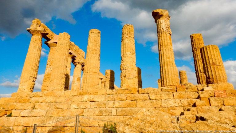 Group of weathered Doric columns of varying heights rising from a golden stone platform beneath blue sky.