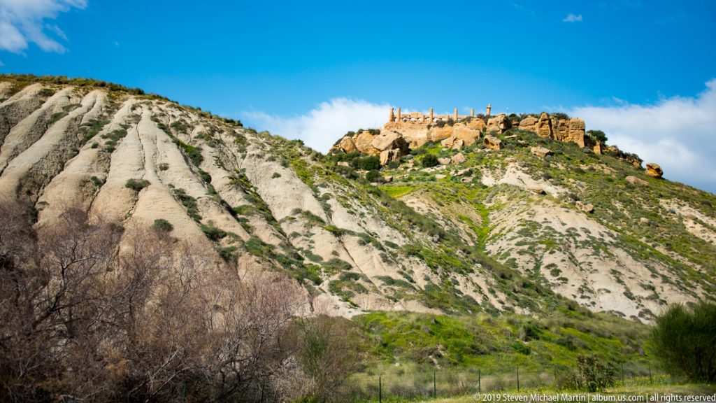 Distant ancient columns and masonry crowning a steep eroded pale ridge beneath a vivid blue sky.