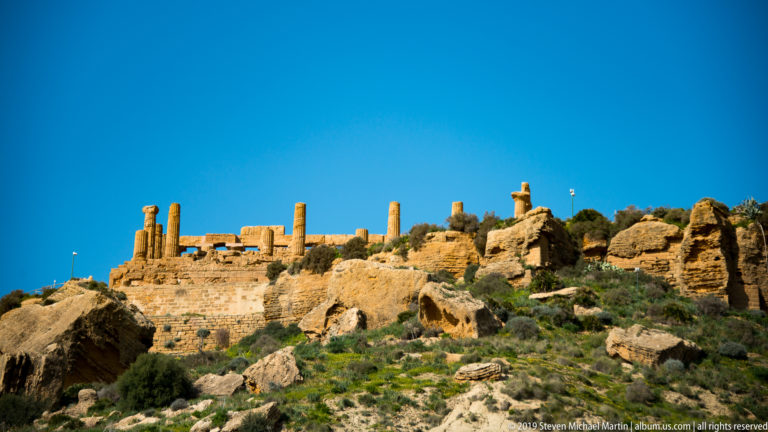 Ancient columns and golden masonry lining the top of a rocky green ridge beneath a deep blue sky.