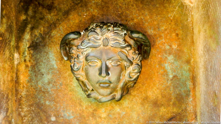 Small classical-style female face ornament mounted on a mottled gold, green, and brown patinated metal surface.