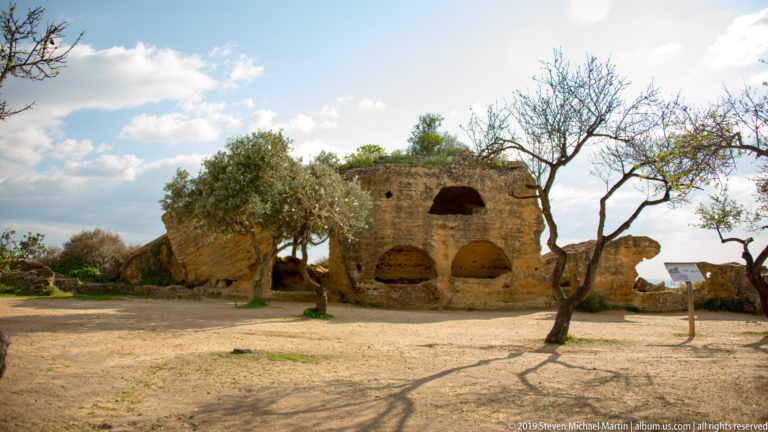 Weathered golden rock and masonry with several arched recesses beneath sparse trees in the archaeological park.