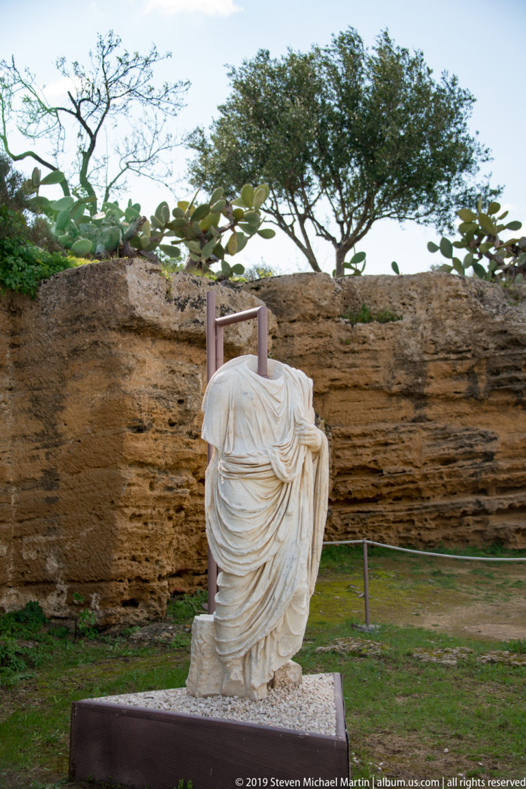 Headless pale draped statue on a base before a layered rock wall, cactus, and trees.
