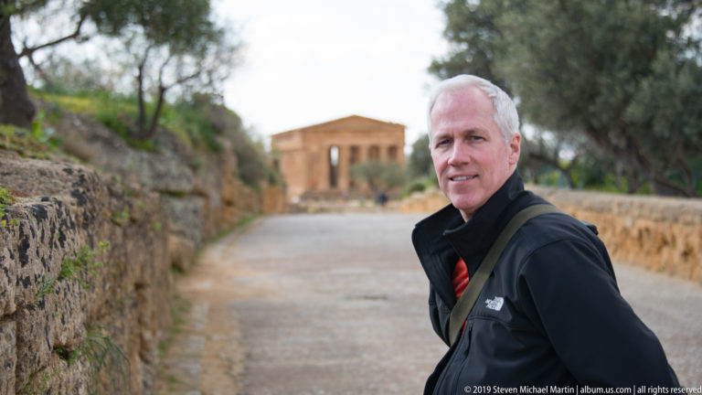 Man in a black jacket on a stone-lined path with the Temple of Concordia softly blurred behind.
