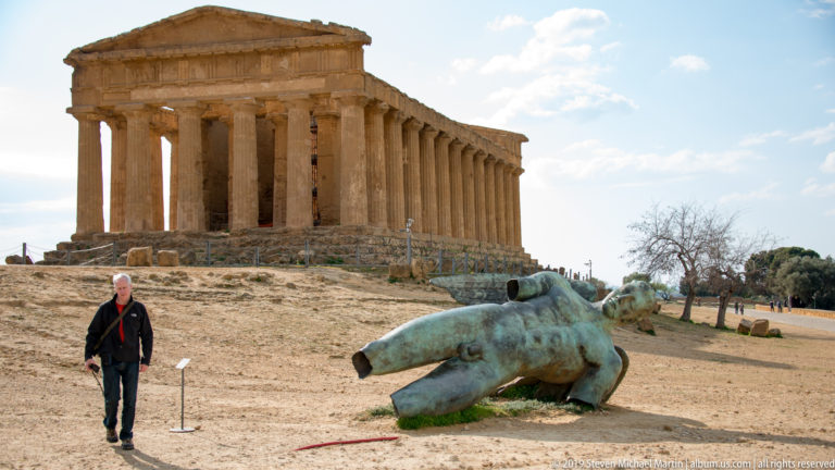 Visitor beside a reclining bronze figure on open ground with the Temple of Concordia rising behind.