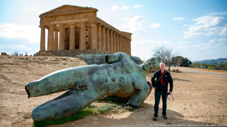 Visitor standing beside a large green-patinated reclining bronze figure with the Temple of Concordia behind.