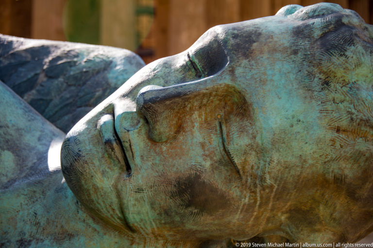 Close detail of a large patinated bronze face lying sideways, showing a closed eye, nose, lips, and textured surface.