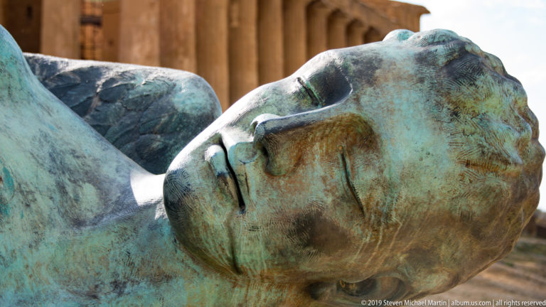 Large patinated bronze head lying on its side with closed eyes, with golden temple columns softly visible behind.
