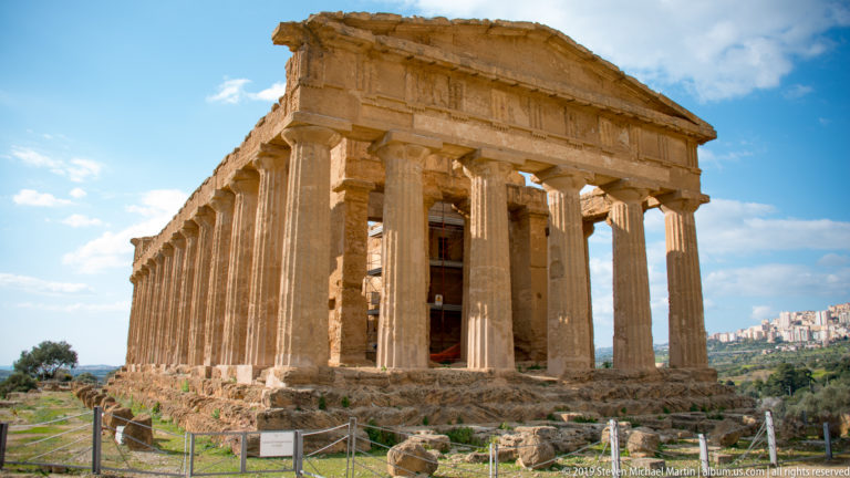 Three-quarter view of the Temple of Concordia with intact pediment and colonnade behind visitor barriers and a small sign.