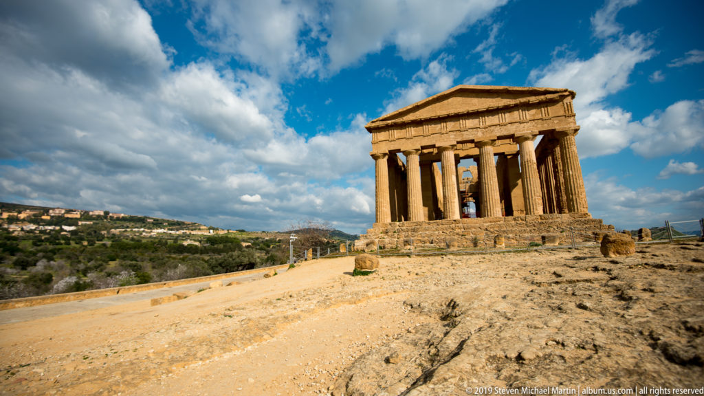 Temple of Concordia standing on a rocky rise at right, approached by pale ground beneath large white clouds.