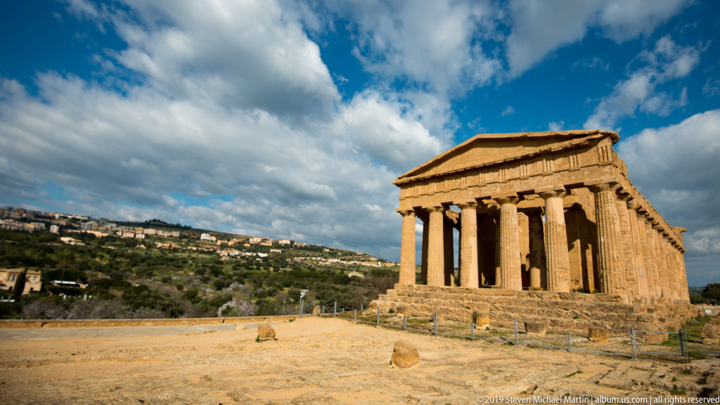 Wide view of the Temple of Concordia amid pale paths and low vegetation beneath a dramatic blue and cloudy sky.