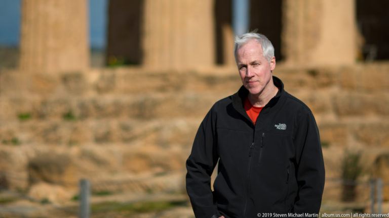 Man in a black jacket standing before the softly blurred stone platform and columns of the Temple of Concordia.