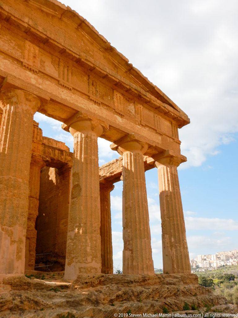 Low oblique view through Doric columns toward the Temple of Concordia's inner walls, with distant city visible beyond.
