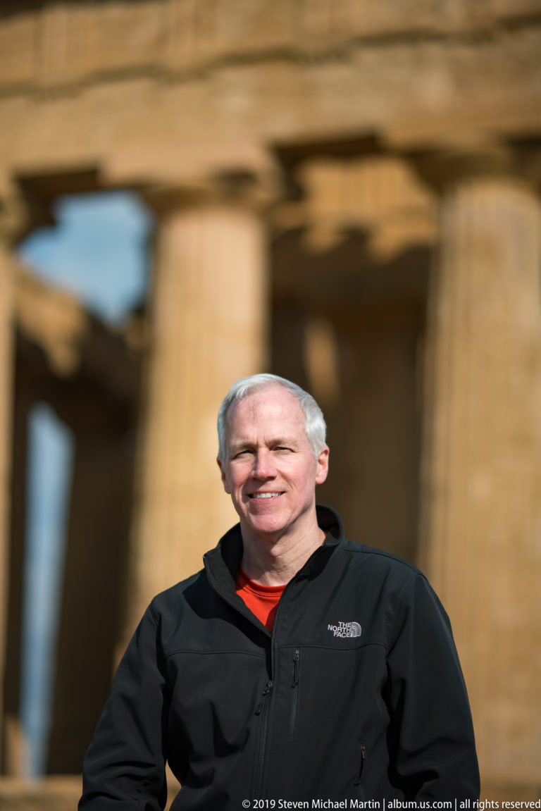 Smiling man in a black jacket standing before softly focused golden columns and entablature of a Greek temple.