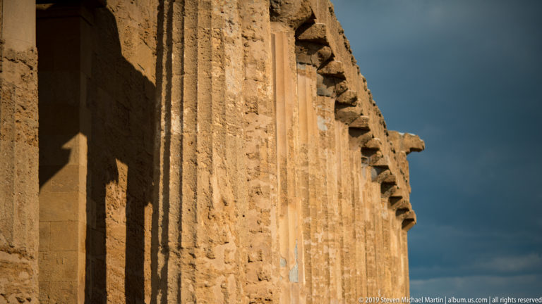 Sunlit weathered stone wall and projecting architectural blocks along the side of the Temple of Concordia beneath dark clouds.