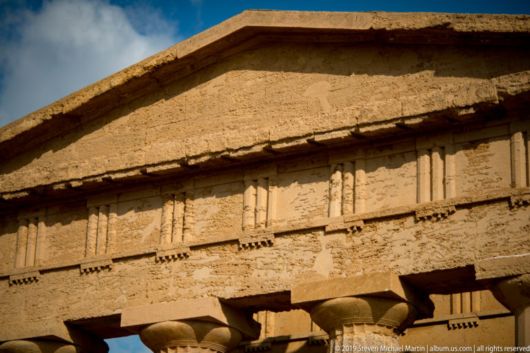 Close view of the golden stone pediment, cornice, and Doric triglyph-and-metope frieze above partial column capitals.