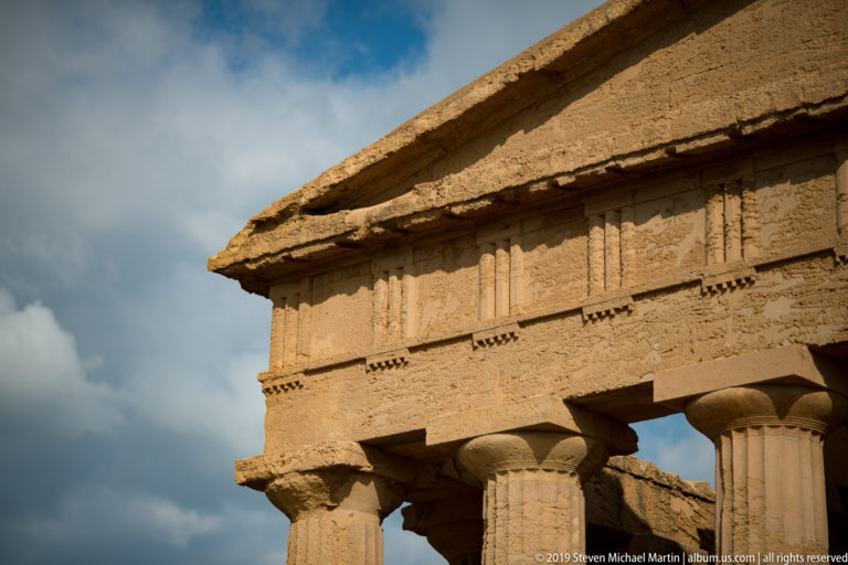 Oblique close view of the Temple of Concordia pediment, Doric frieze, cornice, and column capitals under a cloudy sky.