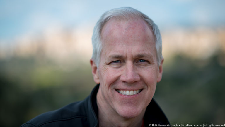 Close portrait of a smiling gray-haired man with a softly blurred green and golden landscape behind him.
