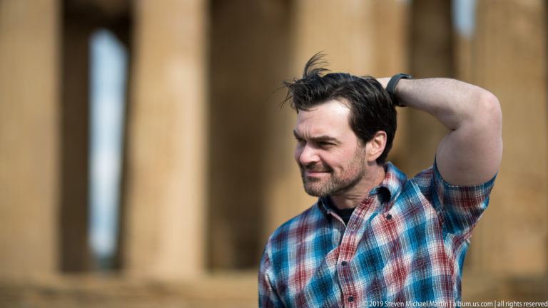 Man in a plaid shirt posing with one hand behind his head before blurred golden Doric columns.
