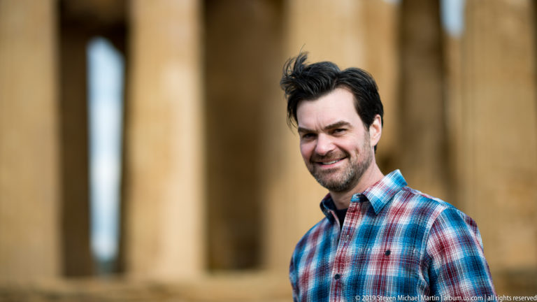 Man in a plaid shirt standing before softly blurred golden temple columns, with wind lifting his hair.