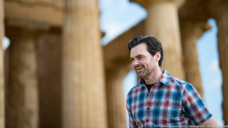 Smiling man in a plaid shirt beside a softly focused row of golden Doric columns and shadowed openings.