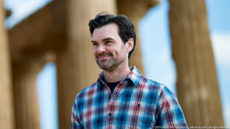 Man in a plaid shirt standing before softly blurred golden Doric columns at the Temple of Concordia.