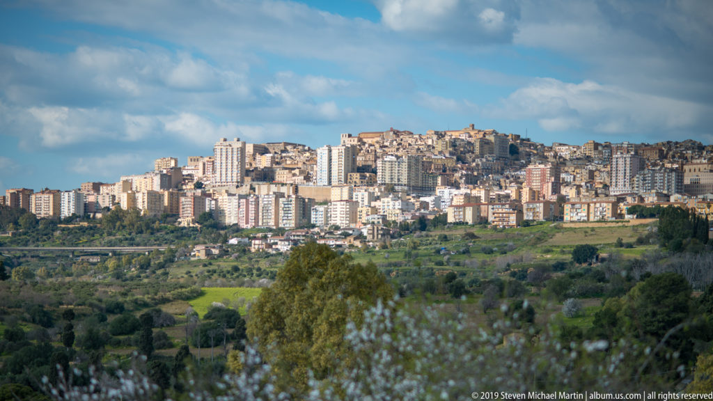 Modern Agrigento densely covering a hilltop ridge above green fields and trees under a partly cloudy sky.