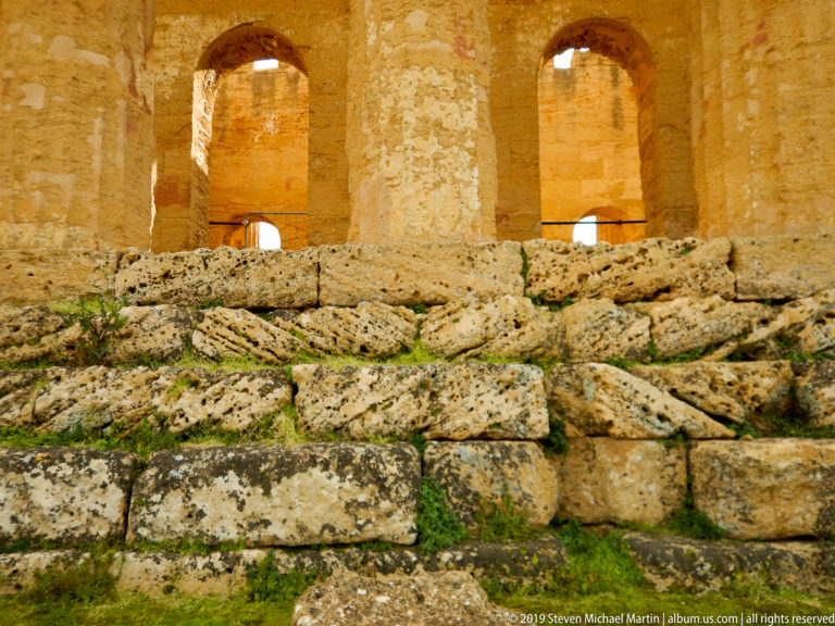 Golden masonry wall with tall arched openings above rough weathered foundation blocks and patches of grass.