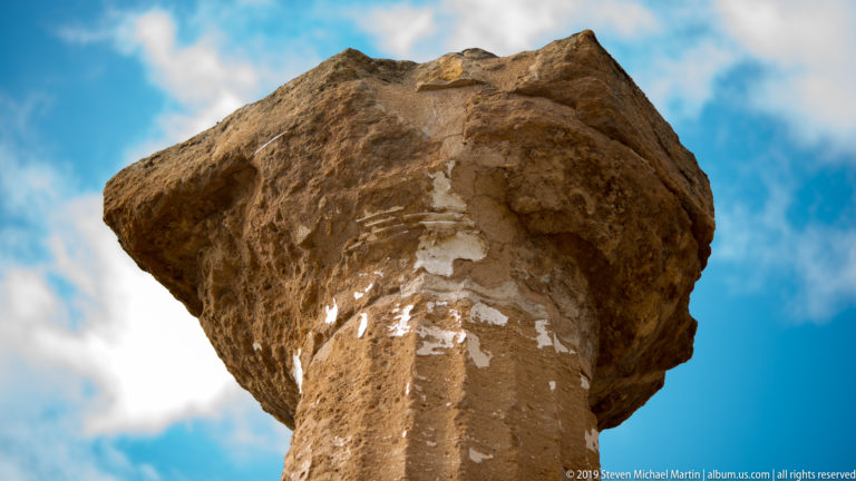 Low-angle close view of a weathered fluted Doric column and eroded capital against blue sky and clouds.