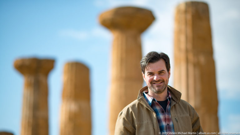 Man standing in front of several softly focused weathered Doric columns and capitals under a pale sky.