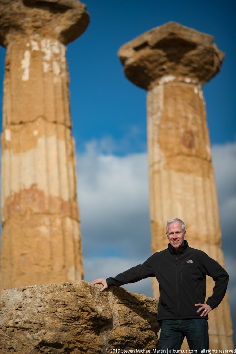 Man posing beside low ruins beneath two massive Doric columns with broad capitals and a darkening sky behind.