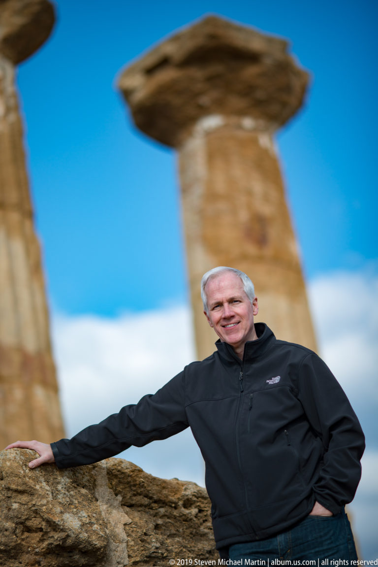 Man leaning on a stone wall with massive weathered Doric columns rising softly out of focus behind him.