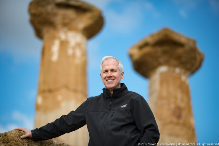 Man in a black jacket standing before two softly focused weathered Doric columns and capitals.