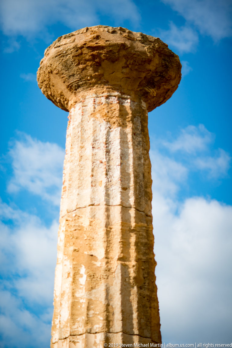 Close upward view of a weathered fluted Doric column and broad capital against blue sky and white clouds.